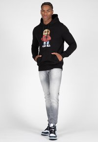Zwarte hoodie met een kleurrijk grafisch ontwerp op de voorkant, gecombineerd met lichtgrijze jeans en zwart-witte sneakers. Beschikt over frontzakken.