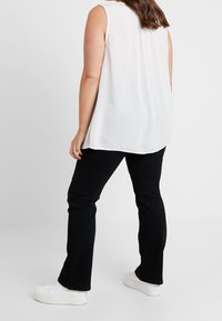 Top bianco senza maniche con orlo arrotondato, abbinato a jeans neri a zampa d'elefante e sneakers bianche con plateau, visto da dietro.