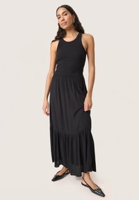 Sort ribbet tanktop parret med en svævende sort maxiskirt med en lagdelt flæsekant. Modellen står i spidse sorte sko.