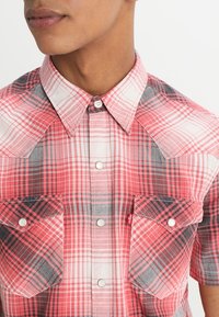 Czerwona i czarna kraciasta koszula z krótkim rękawem i kołnierzykiem na guziki, wyposażona w dwie kieszenie na piersi oraz tkaninę o fakturze.