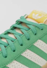 Sneaker verde in suede con strisce bianche, dettagli texturizzati e lacci verdi; presenta una linguetta con marchio giallo e bianco.