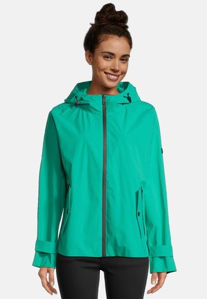 Femme souriante aux cheveux foncés relevés en chignon, portant une veste à capuche turquoise avec fermeture éclair et un pantalon noir, sur un fond uni.