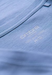 Světle modré bavlněné tričko s kulatým výstřihem, s vyraženým textem uvádějícím značku a detaily o látce v bílé barvě. Hladká textura.