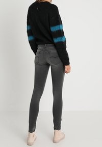 Persona con un suéter negro de felpa con rayas azules en las mangas, jeans ajustados grises y zapatillas de deporte altas en un tono beige claro, de pie de espaldas.