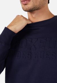Mörkblå sweatshirt i mjukt tyg med en texturerad präglad logotyp, ribbad halsringning och ärmslut, med en avslappnad passform.