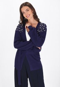 Cardigan en tricot bleu marine avec des accents en perles sur les épaules, manches longues, ceinture à la taille et design ouvert à l'avant. Texture lisse, coupe décontractée.