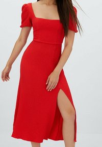 Czerwona sukienka o teksturowanej powierzchni z kwadratowym dekoltem, krótkimi bufkami i rozcięciem do uda, posiadająca dopasowaną górę i zwiewny dół.