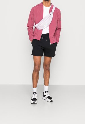 Mężczyzna w różowej kurtce, białej koszulce, czarnych szortach, białych trampkach, białych skarpetach i jasnoróżowej torbie na ramię marki Nike.