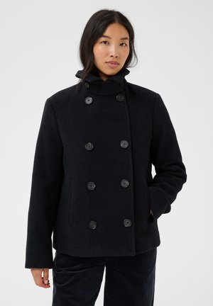 Femme portant un manteau noir croisé à six boutons et col montant, debout avec une main dans sa poche.