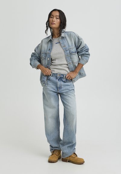 Woman wearing light blue denim jacket, gray t-shirt, baggy jeans, and tan work boots standing against a plain light background.