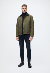 Casaco verde-oliva com fecho de botão e dois bolsos frontais, usado sobre um suéter preto de gola alta, combinado com jeans escuros e botas pretas de tornozelo.