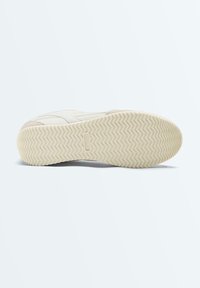 Semelle de chaussure avec motif de bande de roulement en zigzag beige et nom de marque « ONEXXO » embossé au centre, présentée sur un fond clair uni.