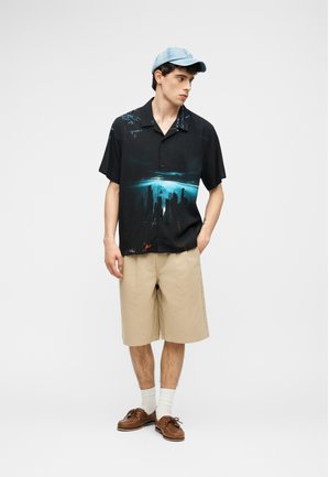 Noor mees, kellel on peas helesinine nokamüts, must lühikeste varrukatega särk linnamaastiku trükiga, beežid põlveni ulatuvad lühikesed püksid, valged sokid ja pruunid kingad.