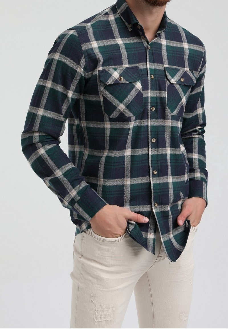 Homme portant une chemise à carreaux vert foncé et bleu marine avec deux poches poitrine, et un pantalon beige, les mains dans les poches.