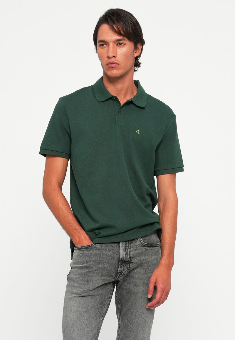 Mężczyzna w ciemnozielonej koszulce polo z krótkim rękawem i małym żółtym logo, w szarych dżinsach, z ręką w kieszeni, stojący na tle jednolitej białej ściany.