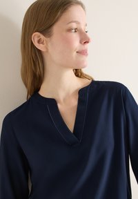 Jeune femme aux cheveux châtain clair portant un haut à manches longues bleu marine avec un col en V en encoche, regardant vers sa droite sur un fond clair.