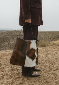 Borsa a tracolla in pelle di mucca marrone e bianca con dettagli in pelle, caratterizzata da una forma morbida e una sola tracolla, indossata accanto a una persona in un abbigliamento scuro.