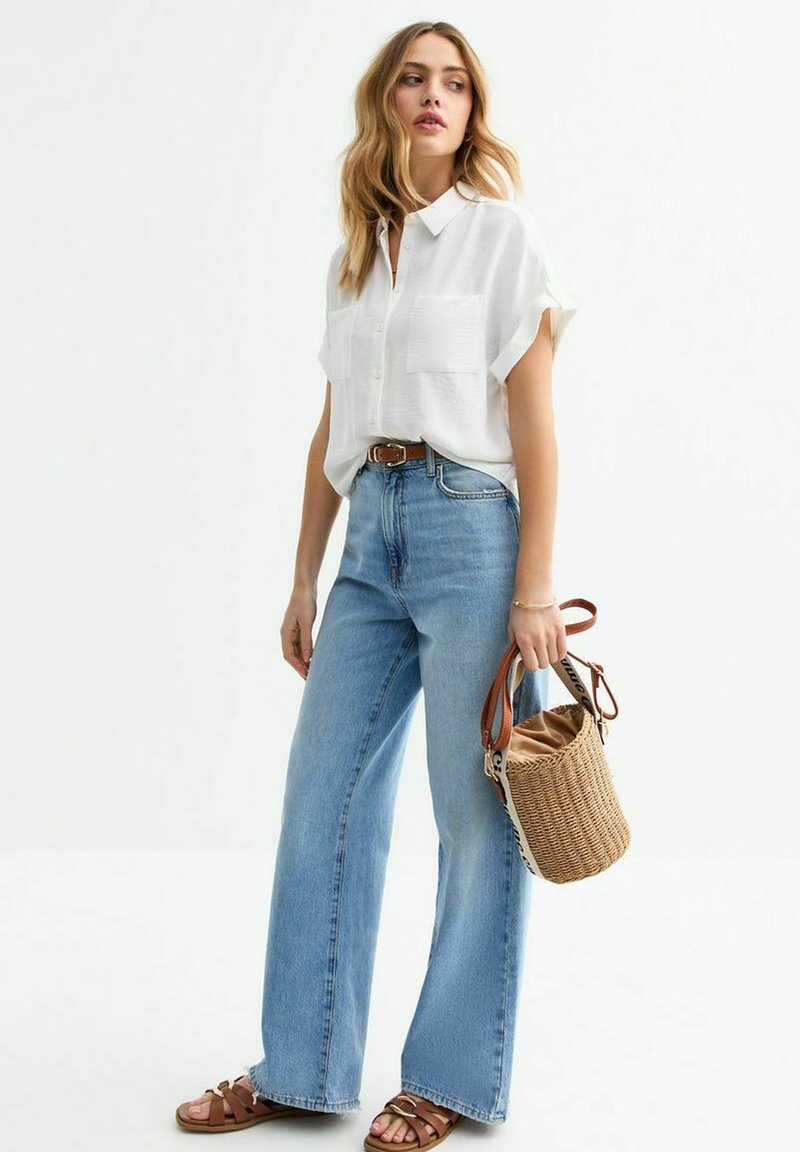 Camicia bianca a maniche corte con due tasche frontali, abbinata a jeans a gamba larga di un leggero blu. Sandali in pelle marrone e una borsa a secchiello intrecciata completano il look.