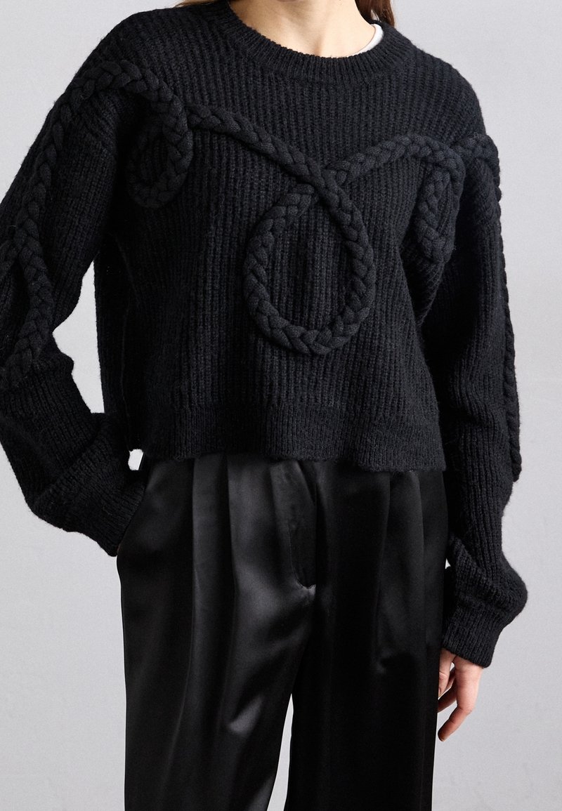 Pull noir en grosse maille avec un large détail de corde tressée en boucle sur le devant et le long des manches, associé à un pantalon en satin noir.