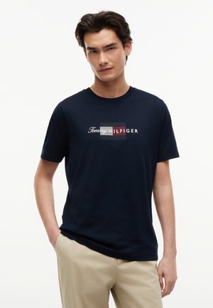 Tânăr purtând un tricou bleumarin cu logo Tommy Hilfiger și pantaloni bej, stând cu o mână în buzunar într-un fundal alb simplu.