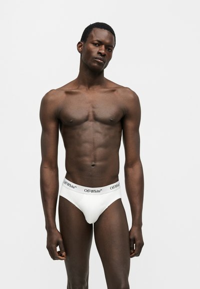 Modèle masculin à la peau foncée portant un slip blanc de la marque Off-White, debout devant un fond gris clair uni.