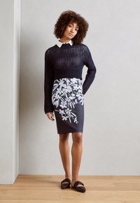 Pull en maille bleu marine au design court, associé à une jupe ajustée ornée de motifs floraux blancs sur fond sombre. Sandales plates brunes.