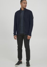 Maglione a zip color navy con colletto alto, polsini a coste e una patch sulla manica sinistra. Abbinato a pantaloni neri aderenti e sneakers nere.
