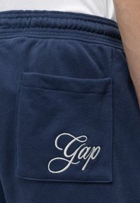 Gros plan sur un pantalon de survêtement bleu marine avec une taille élastique et une poche arrière brodée du mot Gap en écriture blanche.
