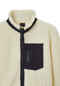 Giacca in pile con un morbido esterno color crema, tasche con zip nere, colletto alto e superficie testurizzata. Chiusura frontale con zip a tutta lunghezza.