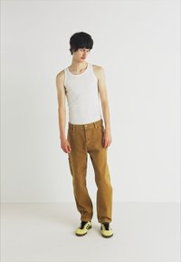 Pantaloni cargo beige con tasche laterali, abbinati a una canotta bianca. Il modello indossa sneakers nere e gialle. Design semplice e senza fronzoli.