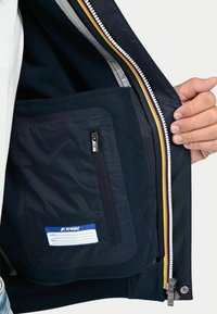 Interno della giacca navy con imbottitura in pile, dettaglio della tasca con zip e un'etichetta blu per nome ed email. Presenta una zip colorata.