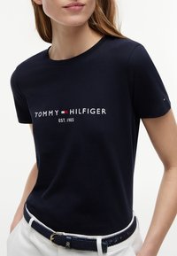 Woman wearing a navy blue Tommy Hilfiger t-shirt with logo and white pants, showing the upper torso and part of the face.