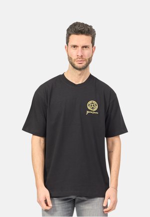 Uomo con capelli corti e scuri e barba, che indossa una t-shirt nera Guess Jeans con logo giallo sul lato sinistro del petto, in piedi davanti a uno sfondo chiaro.