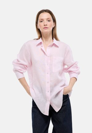 Femme portant une chemise rose pâle à boutons avec les manches retroussées et un pantalon foncé, debout les mains dans les poches devant un fond uni.