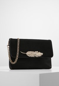 Pochette noire avec bandoulière chaîne en or et fermoir décoratif en forme de plume dorée, posée sur un socle blanc sur un fond uni.