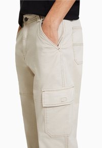 Beige cargo pants with side flap pocket and front slant pocket, worn by person with hand in pocket, paired with black top.