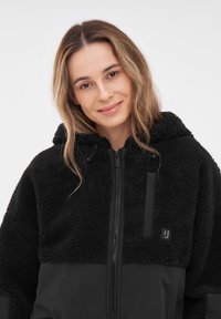 Giacca nera con cappuccio caratterizzata da un texture in pile morbido sulla parte superiore e tessuto liscio nella parte inferiore. Include una zip frontale e un dettaglio con logo.