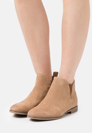 Světle hnědé semišové kotníkové boty s jednoduchým designem, elastickými bočními panely a nízkým dřevěným podpatkem. Povrch je měkký s matným finišem.