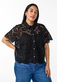 Sort blonde bluse med kortærmer og krave, der har blomster mønstre, knap front og bølgede kanter, kombineret med blå jeans.