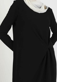 Vestido negro de manga larga con un detalle retorcido en la cintura y una bufanda blanca que cae alrededor del cuello, acentuado por un broche decorativo.