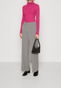 Rosa långärmad polotröja, grå vida byxor och svarta klackstövlar. Svart handväska med en böjd design hålls i ena handen.