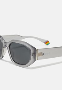 Gafas de sol geométricas grises con lentes oscuros, montura gris transparente, herrajes en tono plateado y un acento a rayas colorido en la patilla.