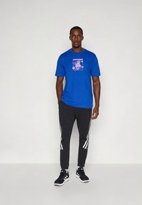 T-shirt de algodão azul com estampa gráfica no peito, combinada com calças de treino pretas com riscas brancas nas laterais e ténis pretos.