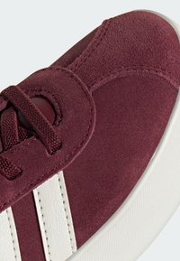 Burgundska suede tenisica s bijelim kožnim prugama. Ima okrugle vezice i teksturiranu gumenu potplatu. Gornji dio ima akcentovane šavove.