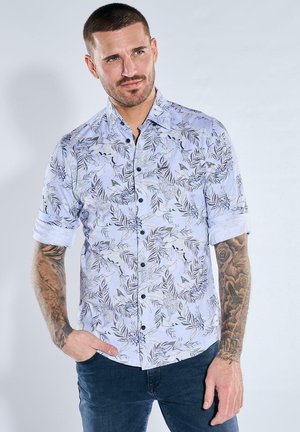 Mann mit kurzen Haaren und Tätowierungen auf beiden Armen, trägt ein hellblaues Hemd mit floralem Muster, aufgekrempelten Ärmeln und dunkle Hosen.