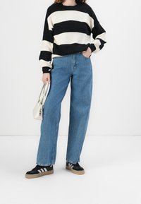 Schwarzer und weiß gestreifter Strickpullover, lockere Passform, kombiniert mit hellblauen weit geschnittenen Jeans. Schwarze Sneakers mit weißen Streifen und einer kleinen weißen Handtasche.