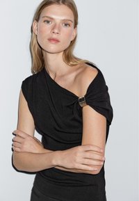 Top nero aderente con un design asimmetrico e un dettaglio dorato sulla spalla. Tessuto morbido, maniche corte e una silhouette pulita.