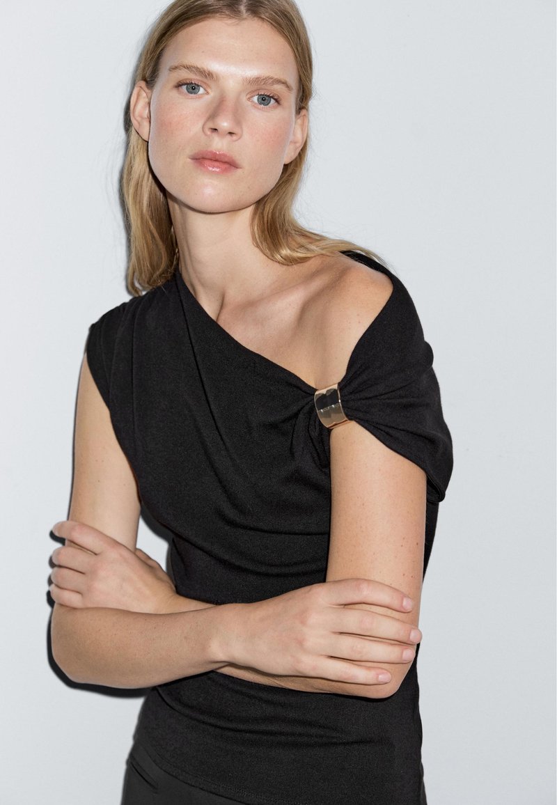 Top nero aderente con un design asimmetrico e un dettaglio dorato sulla spalla. Tessuto morbido, maniche corte e una silhouette pulita.