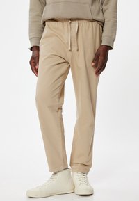 Beige, lette bomuldsblandede bukser med elastisk talje, snørelukning og lige ben-design. Kombineret med hvide sneakers.