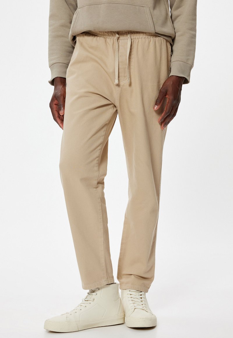Beige, lette bomuldsblandede bukser med elastisk talje, snørelukning og lige ben-design. Kombineret med hvide sneakers.